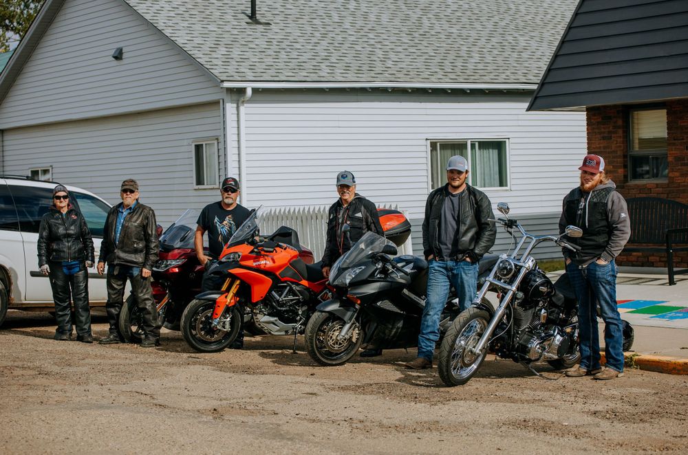 Group of motorcycle enthusiasts gathered with their bikes outside a white house on a cloudy day.