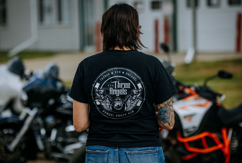 Back view of black t-shirt with circular logo design worn near parked motorcycles.