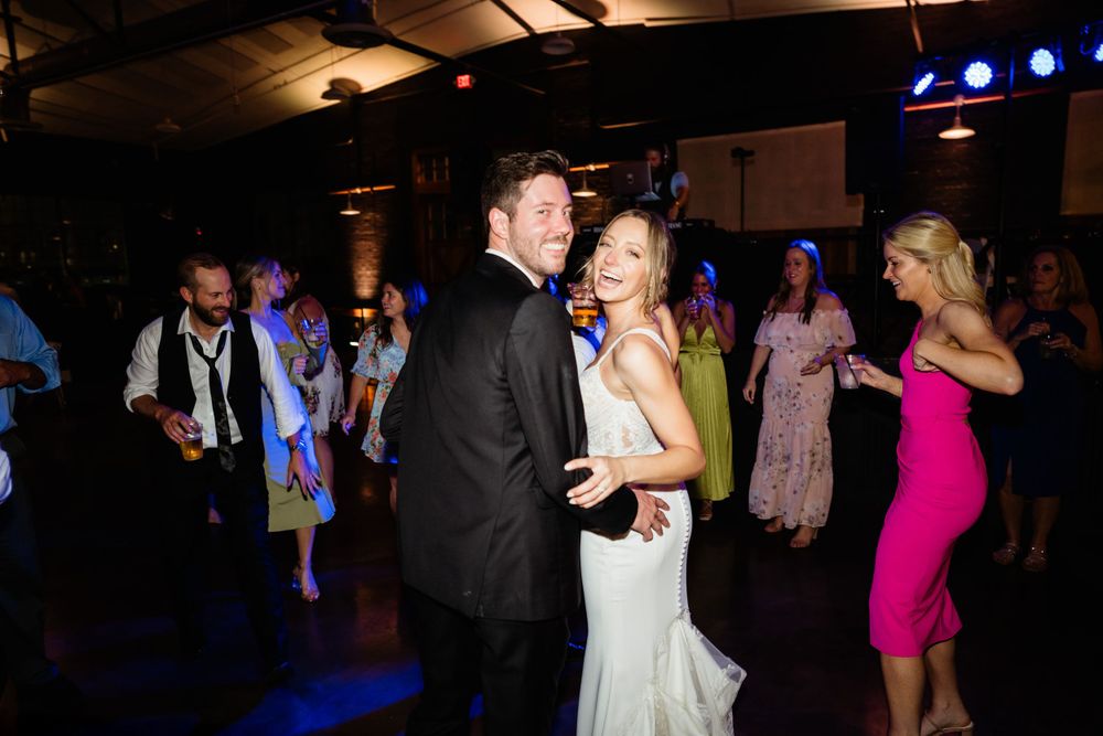 A couple dances at a wedding reception with other guests in formal attire on a dark dance floor with blue lighting.