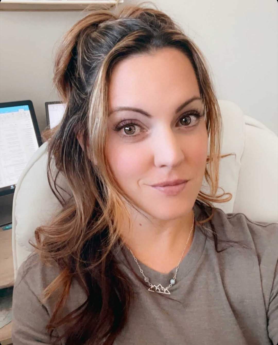A professional selfie showing a woman in casual attire sitting at a desk with a computer in the background.