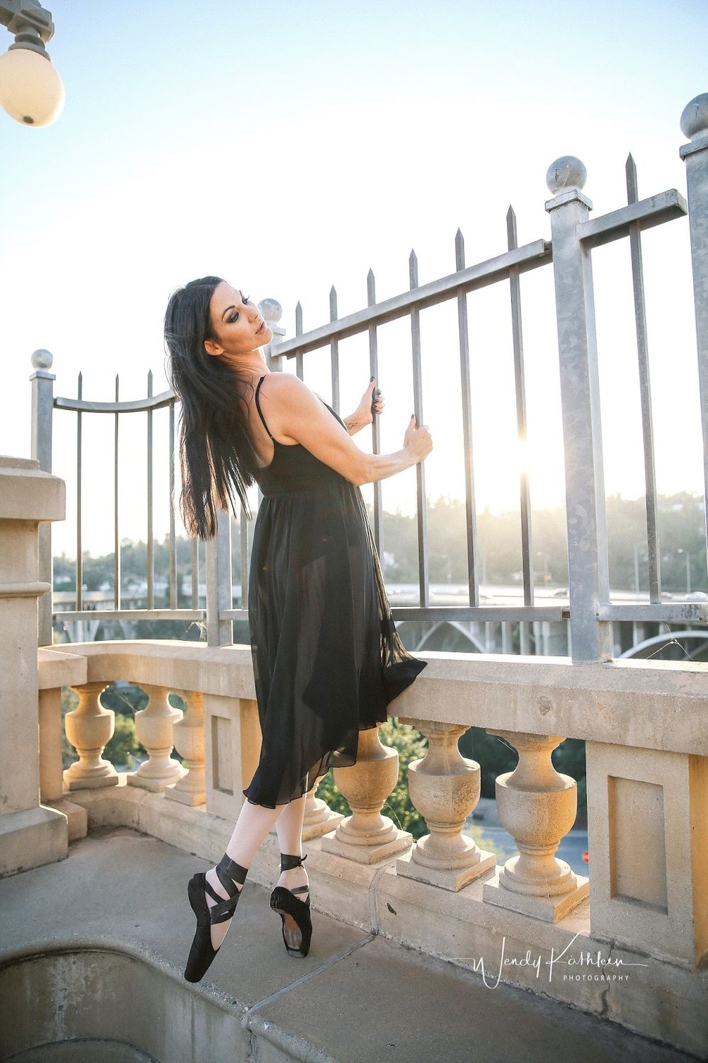 Ballet dancer in black dress poses gracefully on stone balcony with ornate railings at sunset.