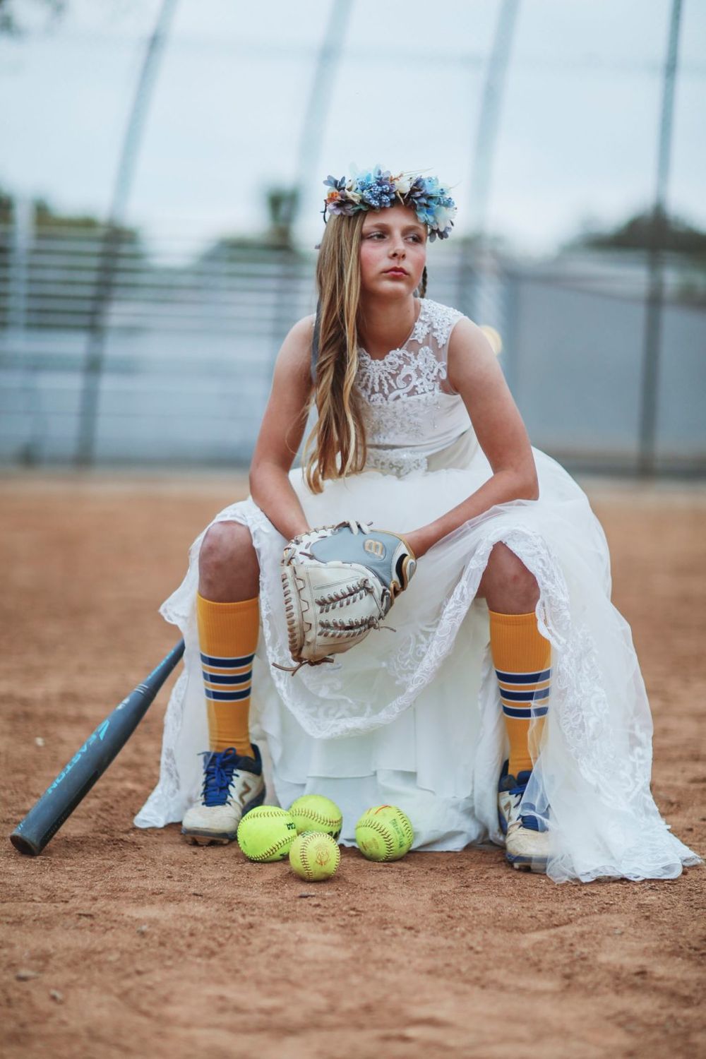 Softball player wearing a white wedding dress and flower crown sits on pitcher's mound with bat and yellow balls.