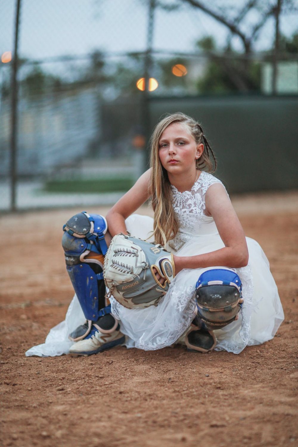 On a baseball field at dusk, a person in a white dress and softball catcher's gear kneels in the dirt.