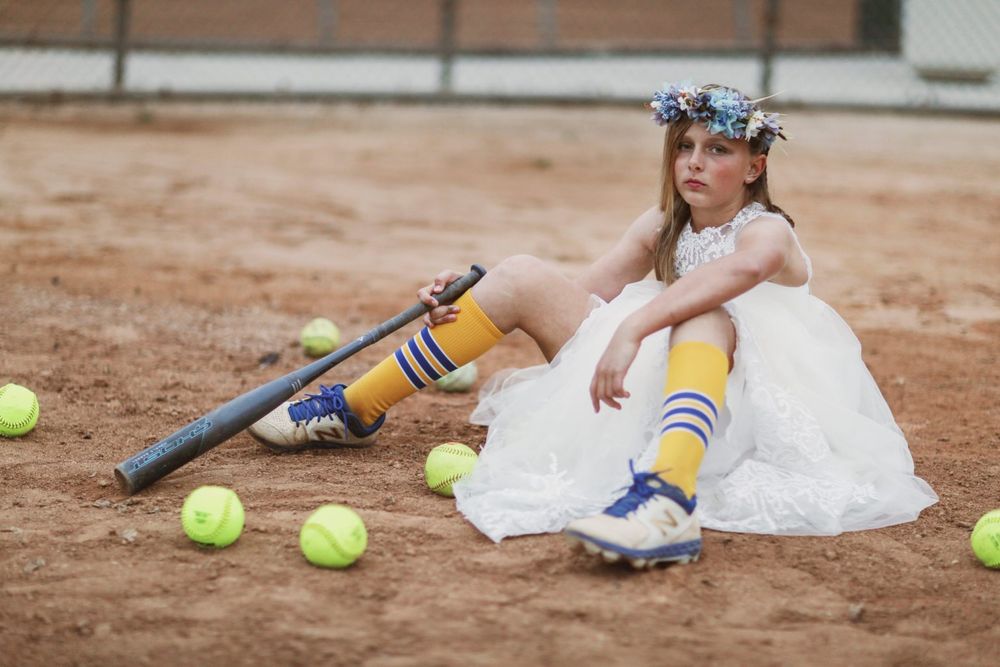 A softball player in a white dress and striped socks poses on a dirt field with scattered tennis balls.