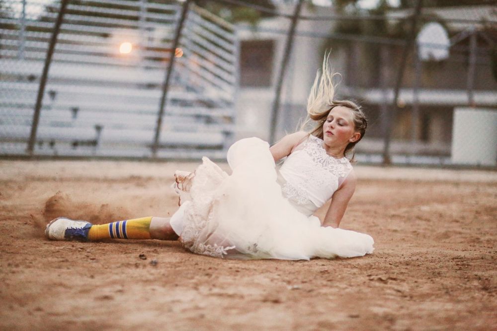 Bride in flowy white dress and athletic socks sits playfully on dirt field at baseball stadium.