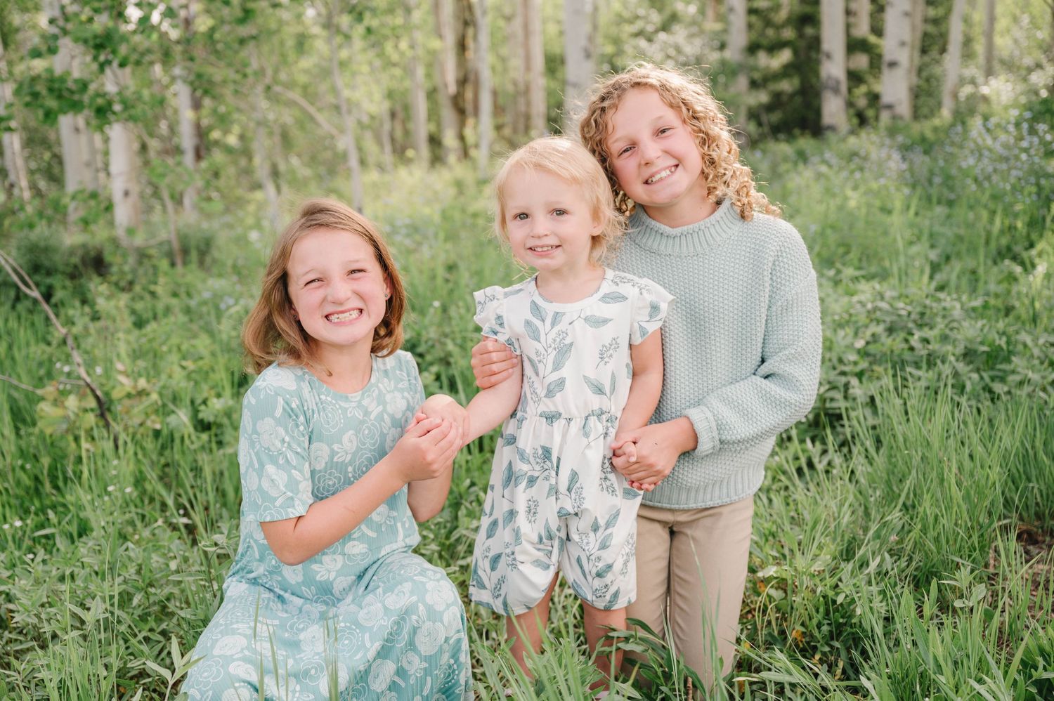 Three children in pastel dresses pose together among aspen trees in a mountain forest setting.