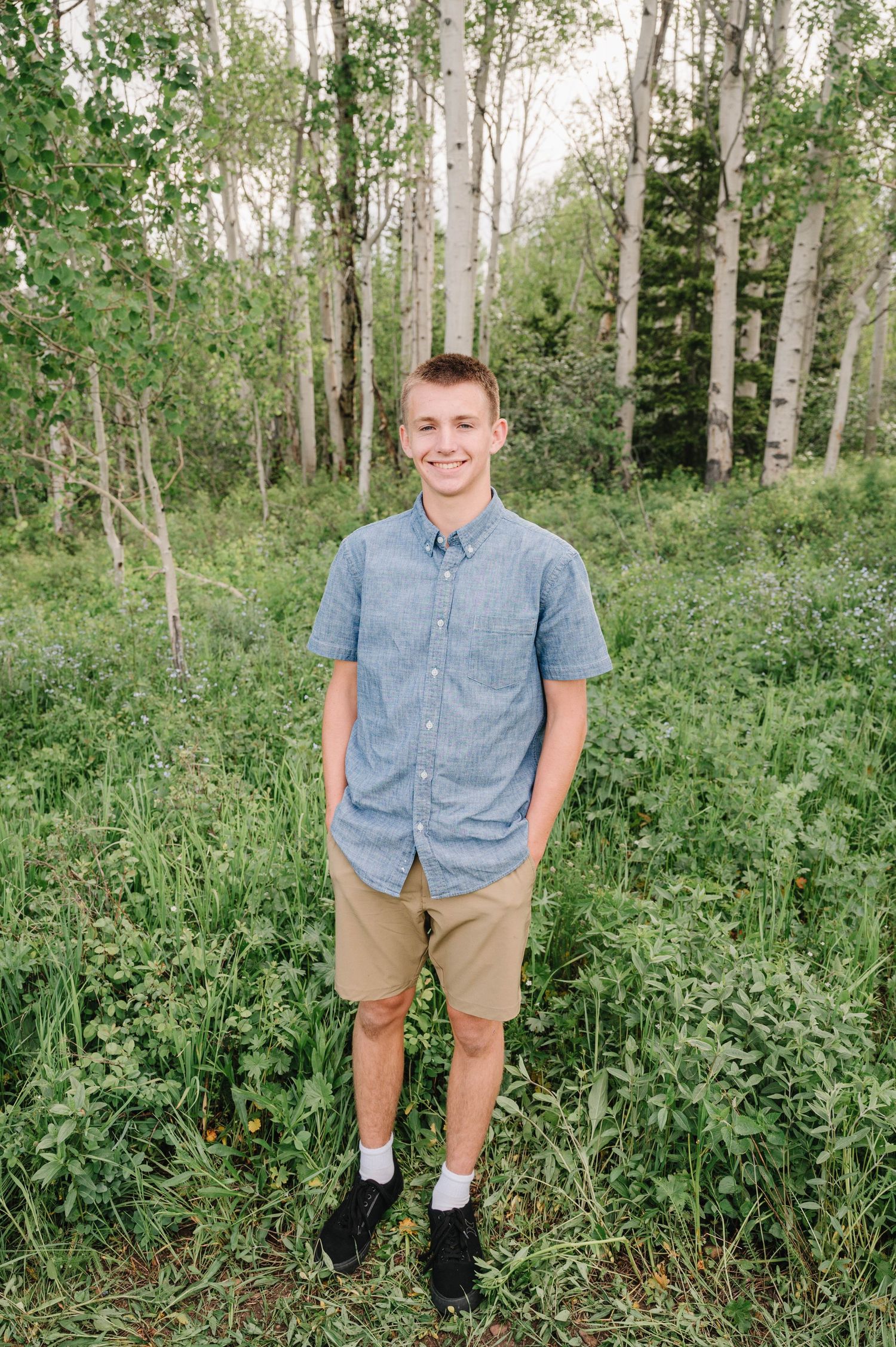 Young person in blue button-down shirt and khaki shorts stands smiling in aspen forest.