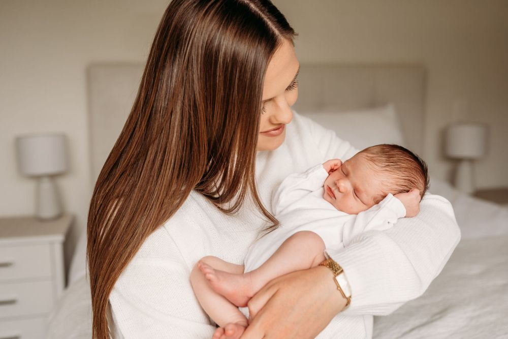 Mother in white sweater cradling sleeping newborn on bed.