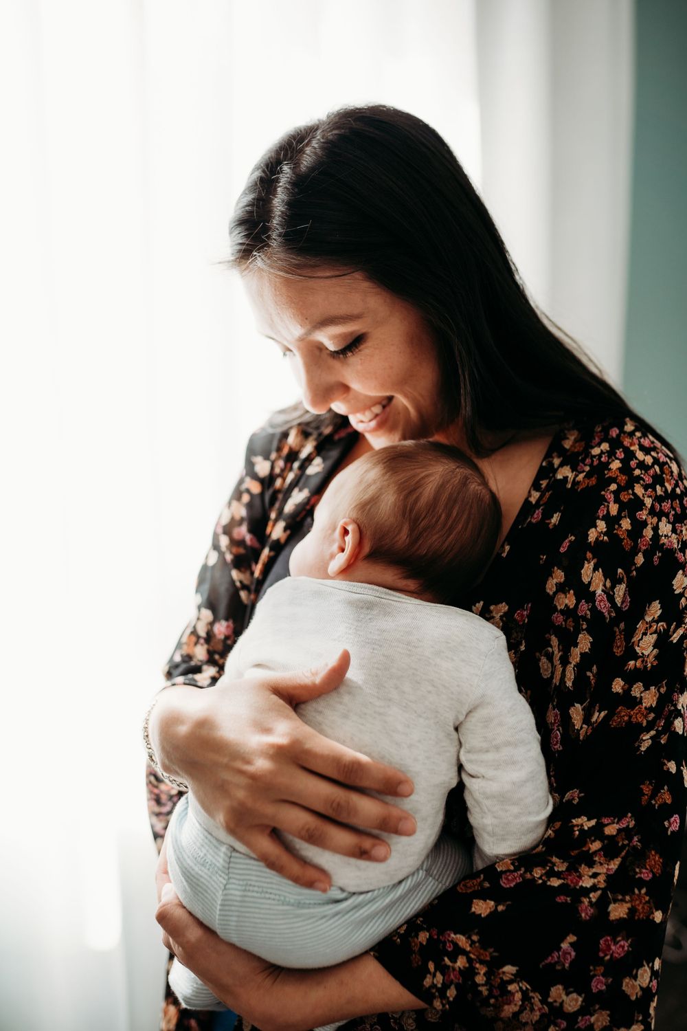 Woman in floral dress holds baby close in tender indoor moment.
