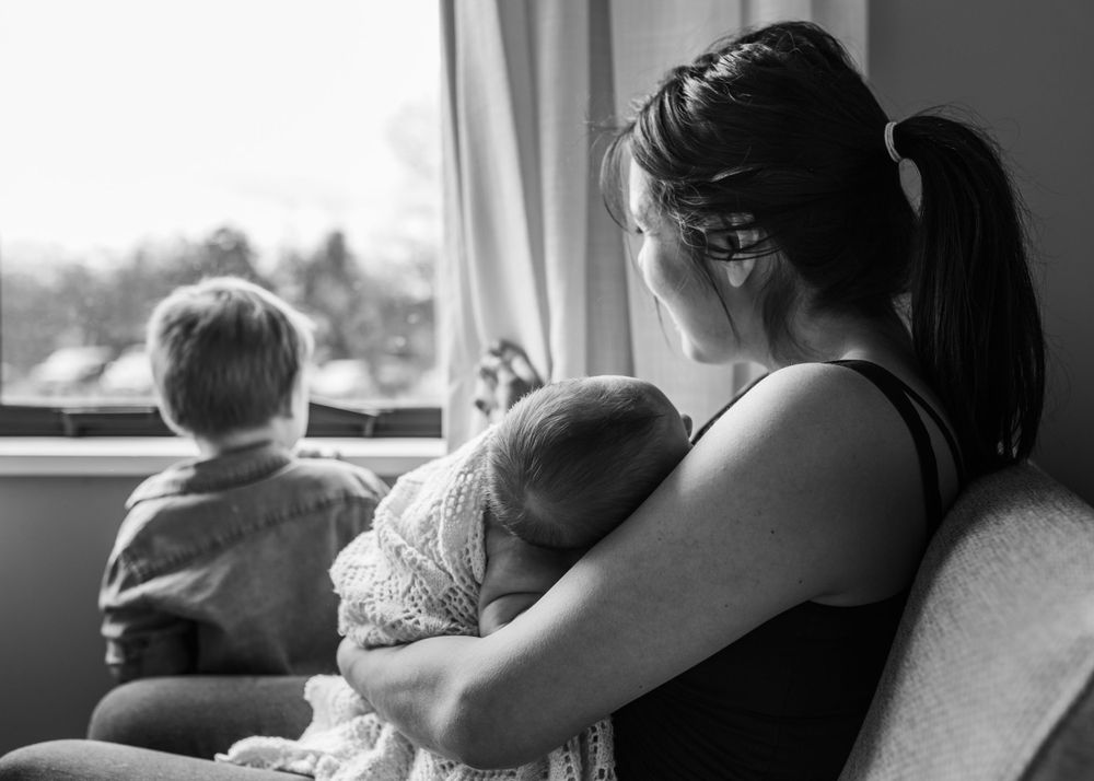 Mother holds sleeping baby while toddler looks out window in black and white photo.