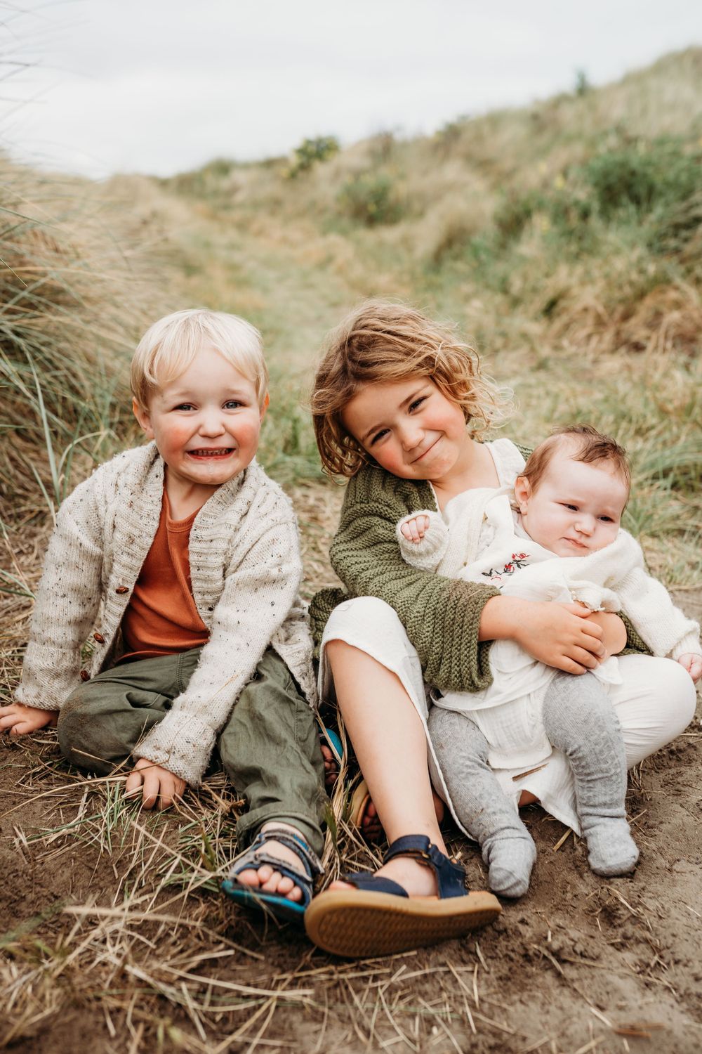 Three young children sit together on a sandy path surrounded by tall grass.