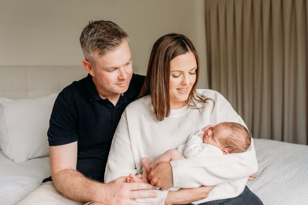 Family of three sitting on bed with newborn baby in white outfit.