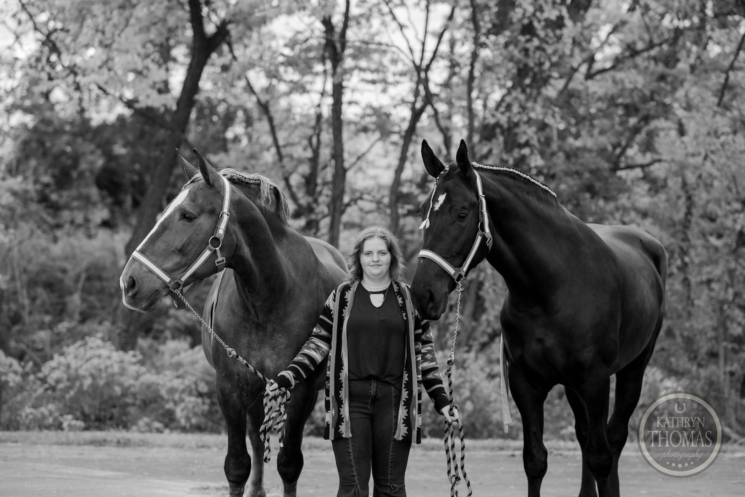 A black and white photo showing two dark horses with bridles being led outdoors in a wooded setting.