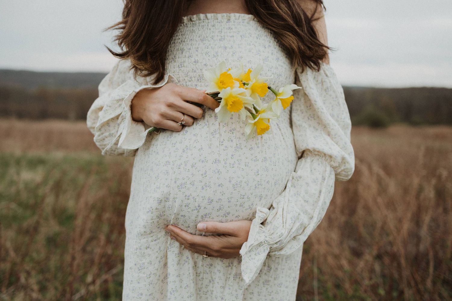 A person in a white lace dress holds yellow daffodils while cradling their pregnant belly.