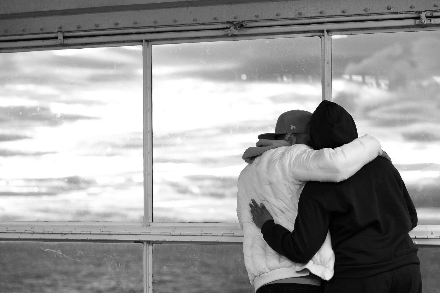 Two silhouettes embrace while looking out window at sunset.