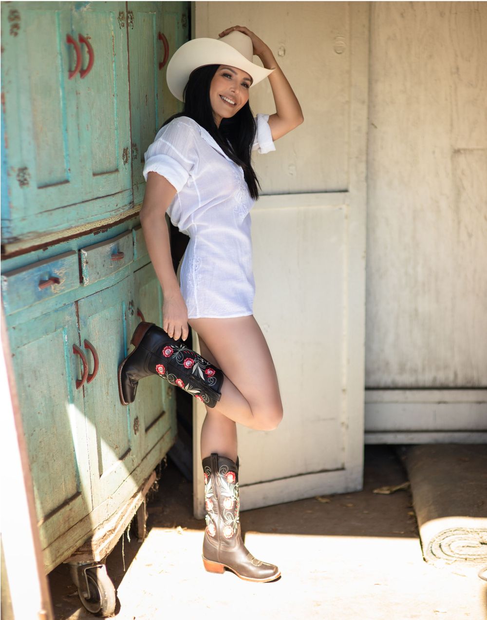 Cowgirl in white outfit and boots poses playfully against a rustic turquoise door.