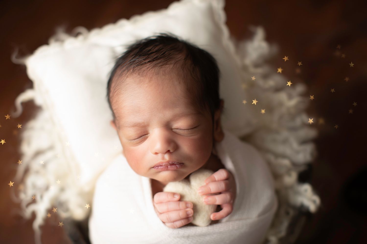 Sleeping newborn baby wrapped in white swaddle on cream pillow with golden star bokeh background.