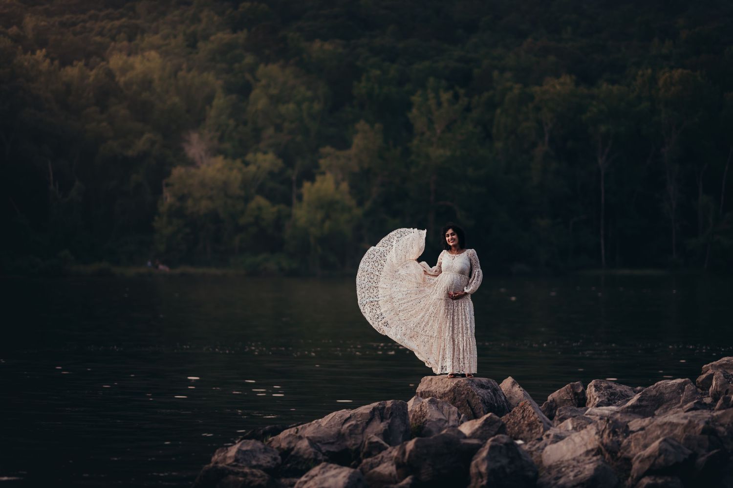 Pregnant mama shows off the flowy skirt of a lace client closet dress while standing on rocks by the river at sunset