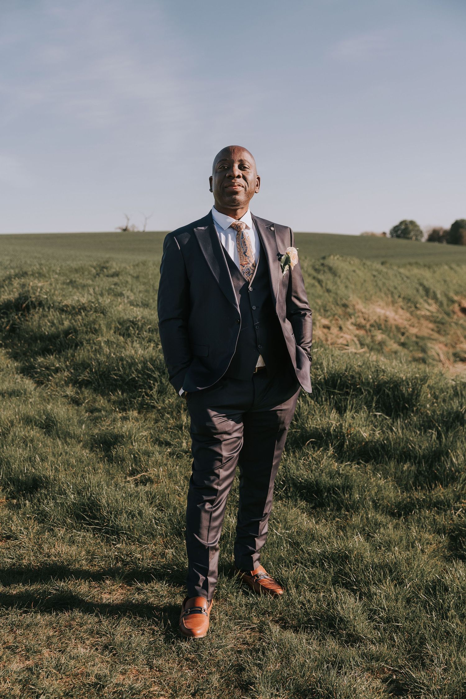 A groom poses in a stylish navy suit and brown shoes on a grassy hillside during golden hour.