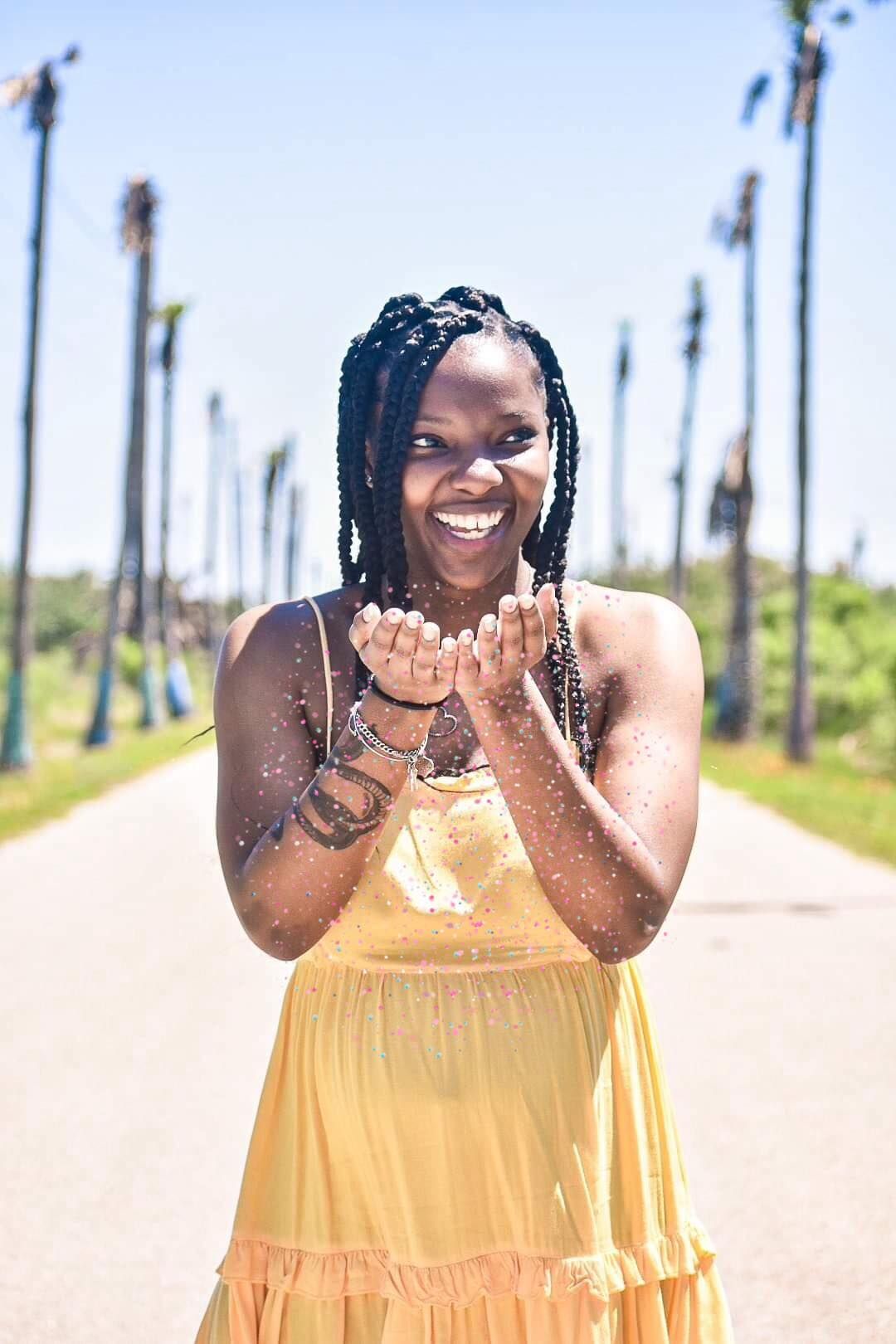 Person in yellow sundress smiling joyfully on palm tree-lined path on sunny day.