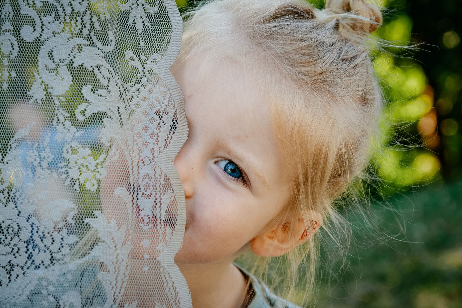 Close-up portrait of a child with blonde hair peeking through delicate white lace curtains.