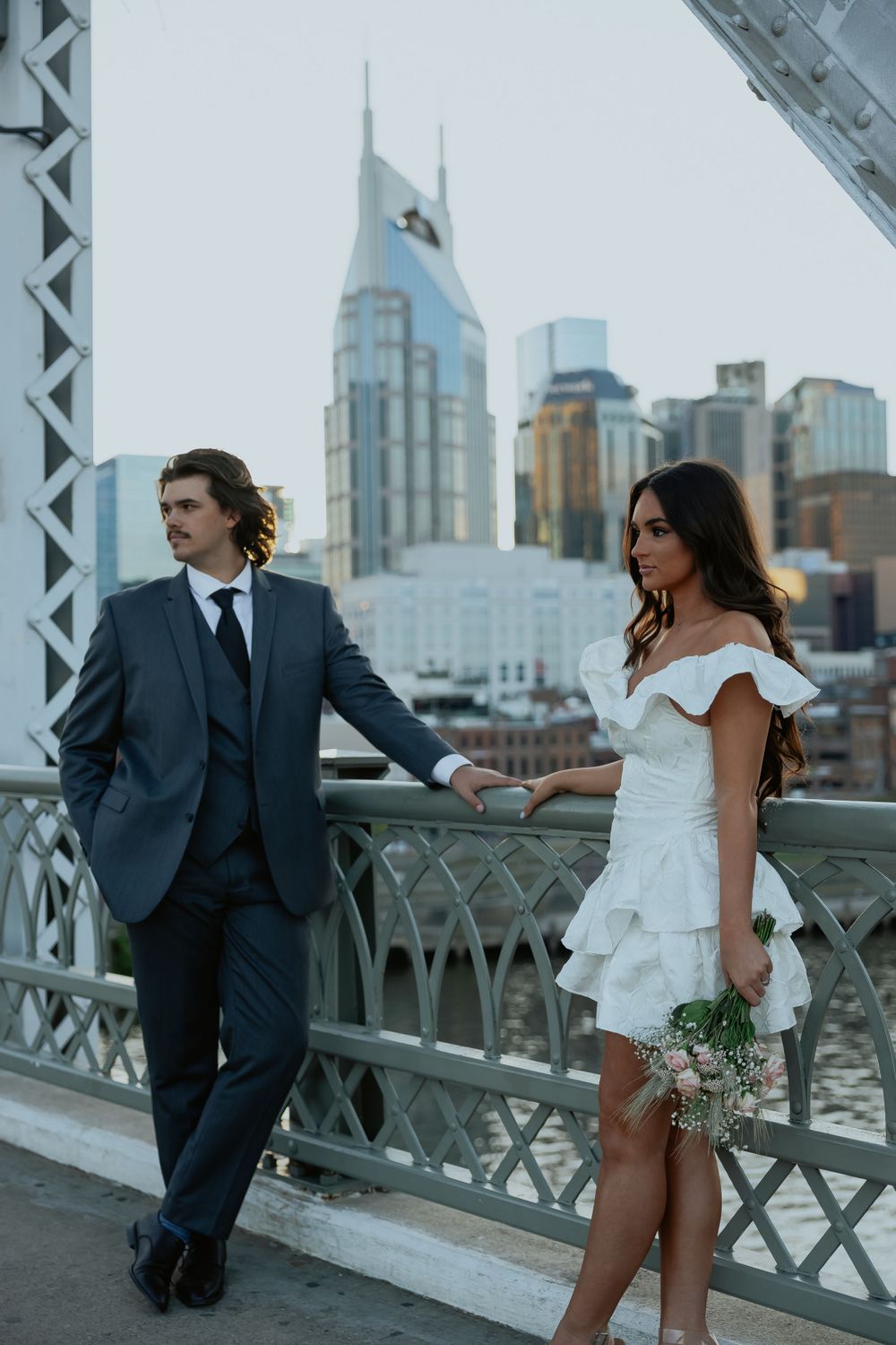 A stylish couple poses on a bridge with Nashville's iconic skyline in the background.