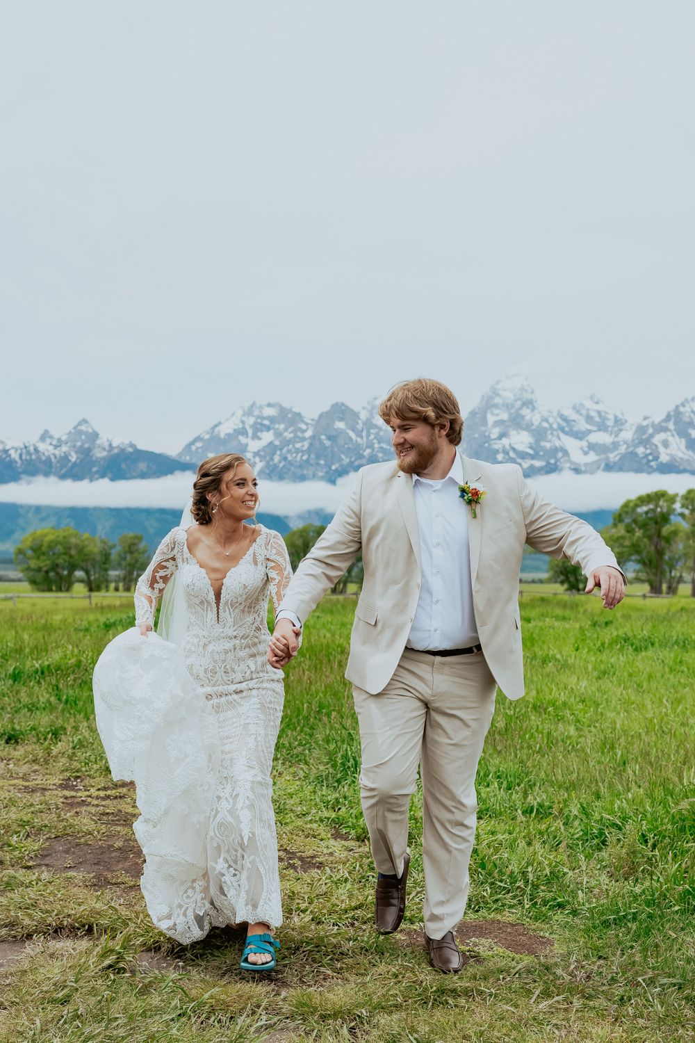 A couple in white and cream attire walking through a lush green field with mountain views.