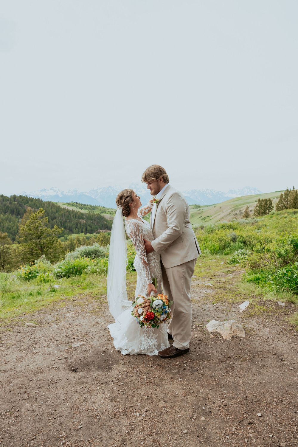 A couple shares an intimate moment on a mountaintop trail while holding a vibrant bouquet.