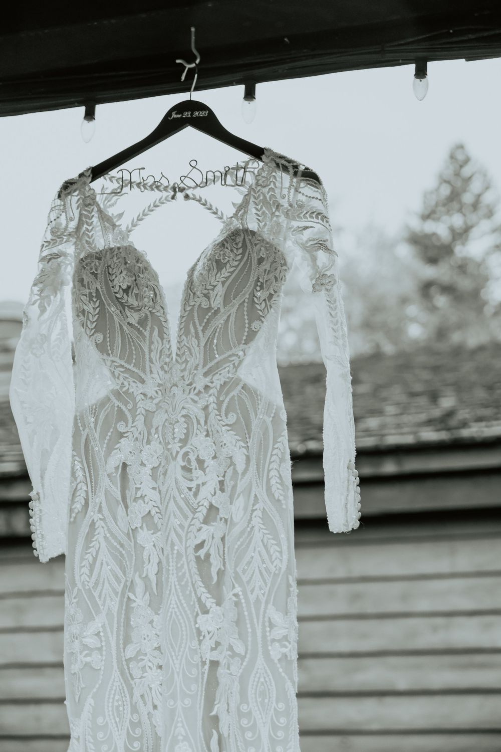 An elegant lace wedding dress with intricate details hangs on display against a rustic background.