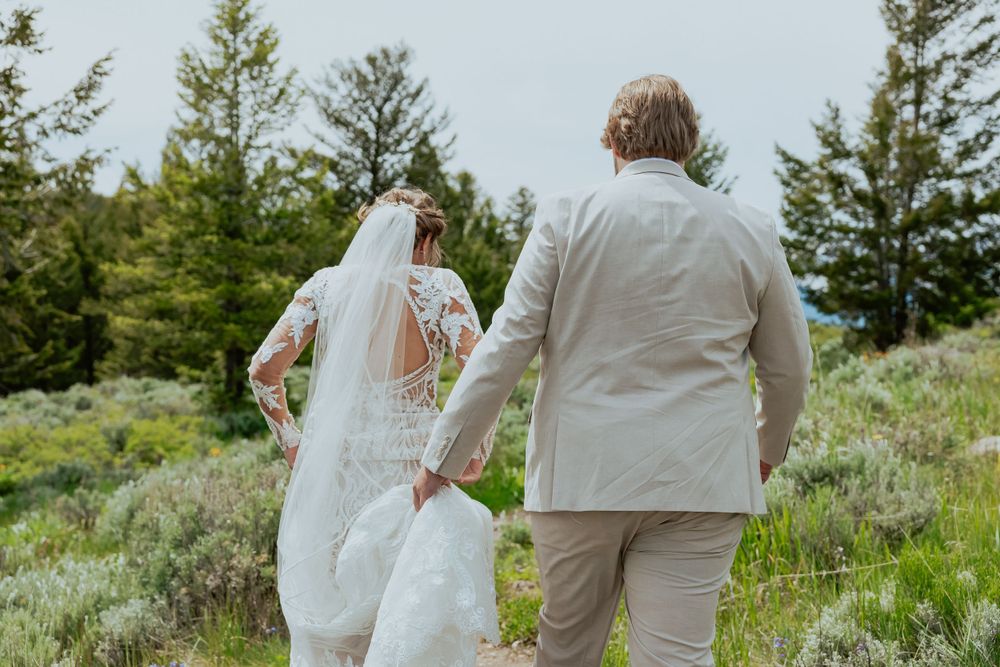 A couple in wedding attire walks hand in hand through a scenic mountain landscape with evergreen trees.