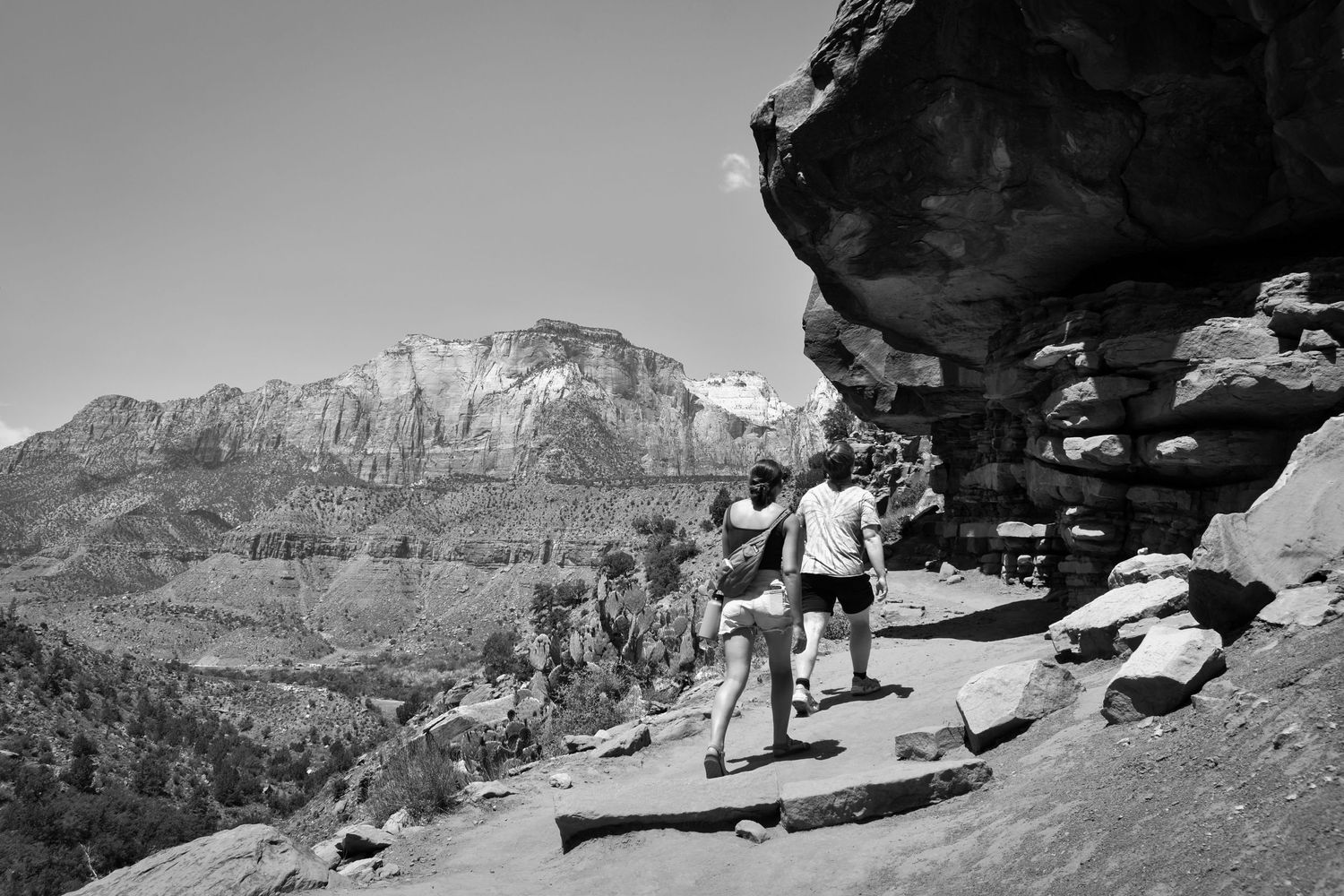 Hikers traverse a narrow mountain trail beneath towering red rock cliffs in a scenic canyon landscape.