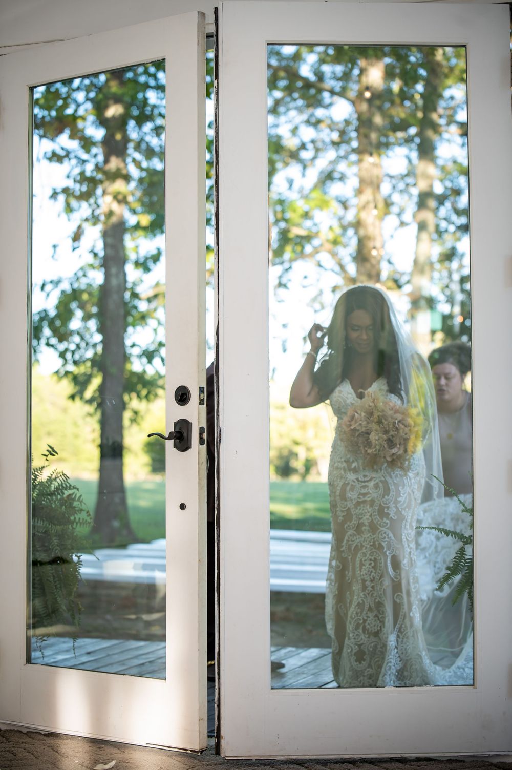 Bride reflected in glass doors while adjusting her veil in natural sunlight.