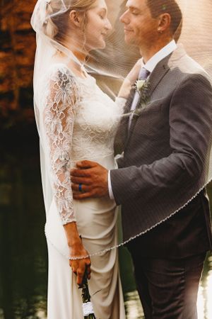 Sunlit wedding portrait through bride's veil as couple shares intimate moment by water.