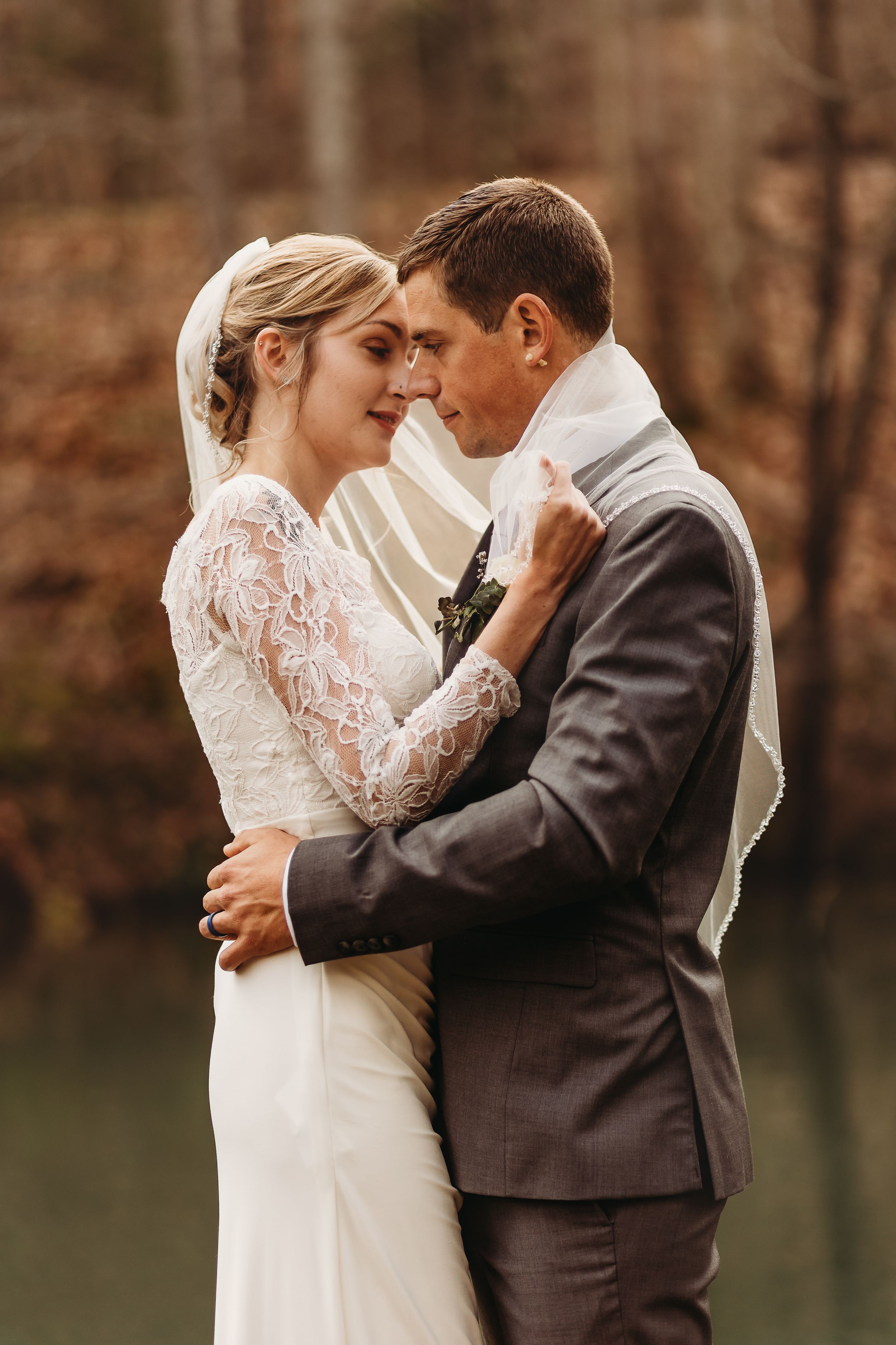Intimate wedding portrait of couple embracing by lake in autumn forest setting.