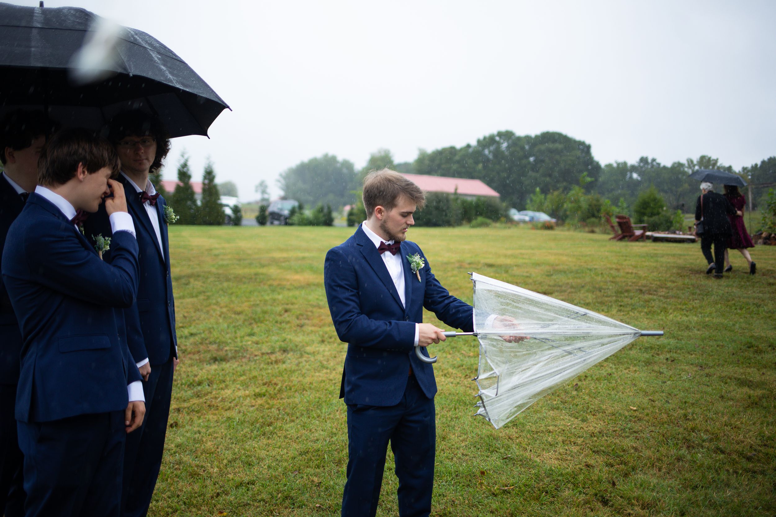 Wedding party members in navy suits stand with umbrellas in a misty field.