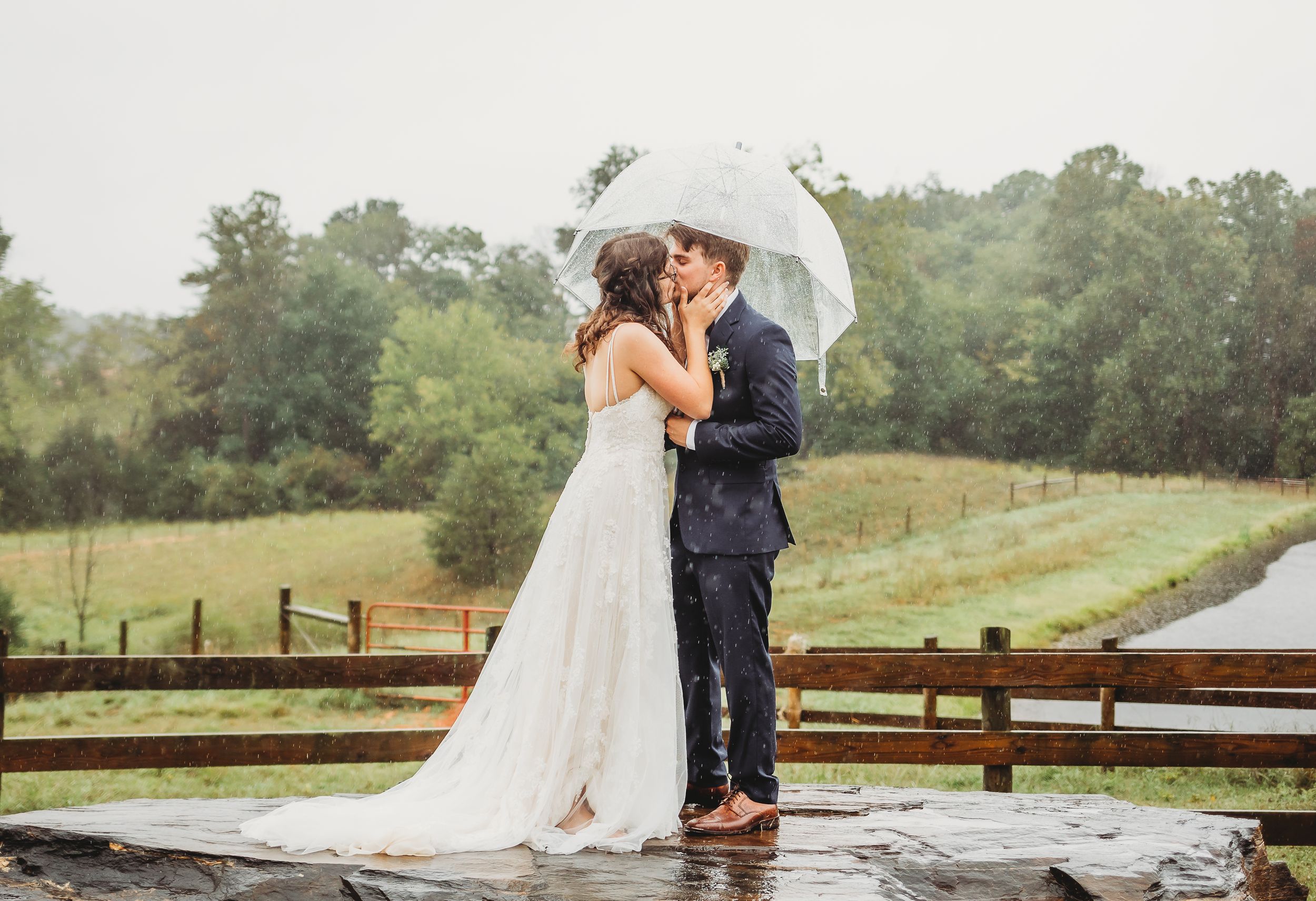Newlyweds share intimate moment under white umbrella in the rain by wooden fence.