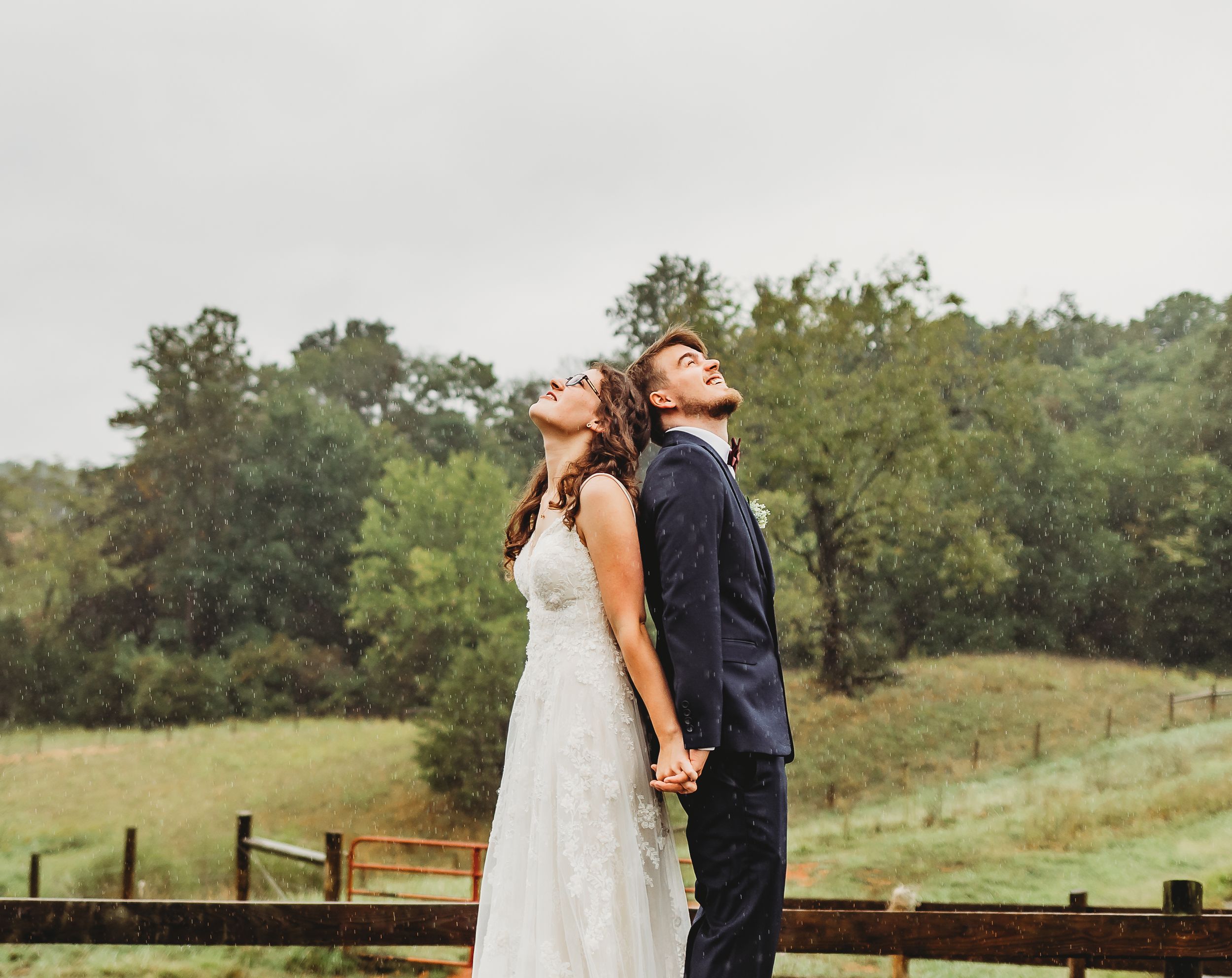 A couple in wedding attire share a romantic moment by a rustic wooden fence on their rainy wedding day.