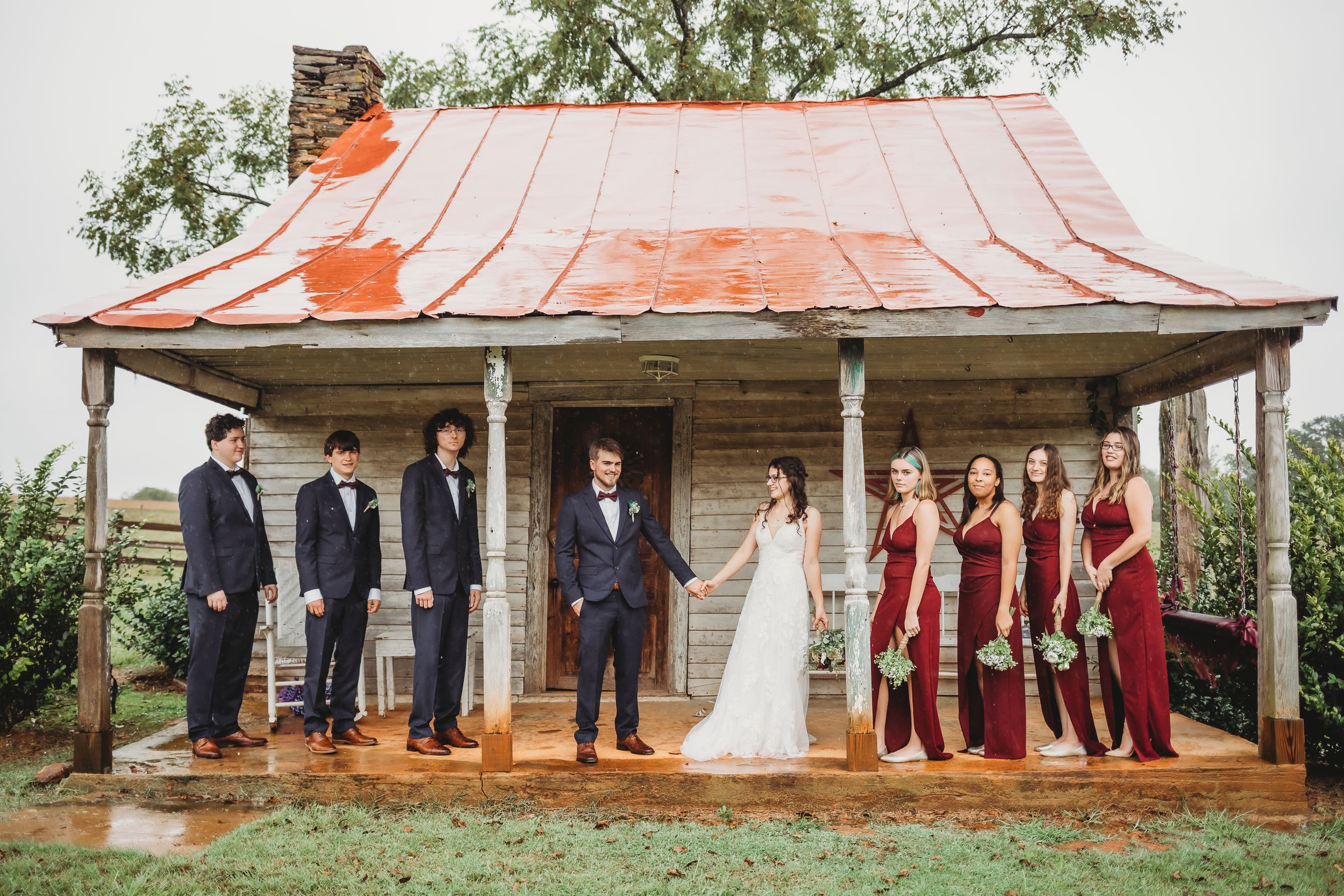 Wedding party poses under rustic wooden pavilion with red metal roof.