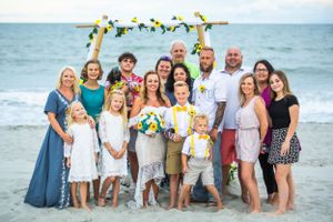 Large family group photo on beach with wedding arch and sunflowers in background.