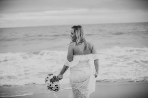 Black and white photo of bride walking on beach holding bouquet.