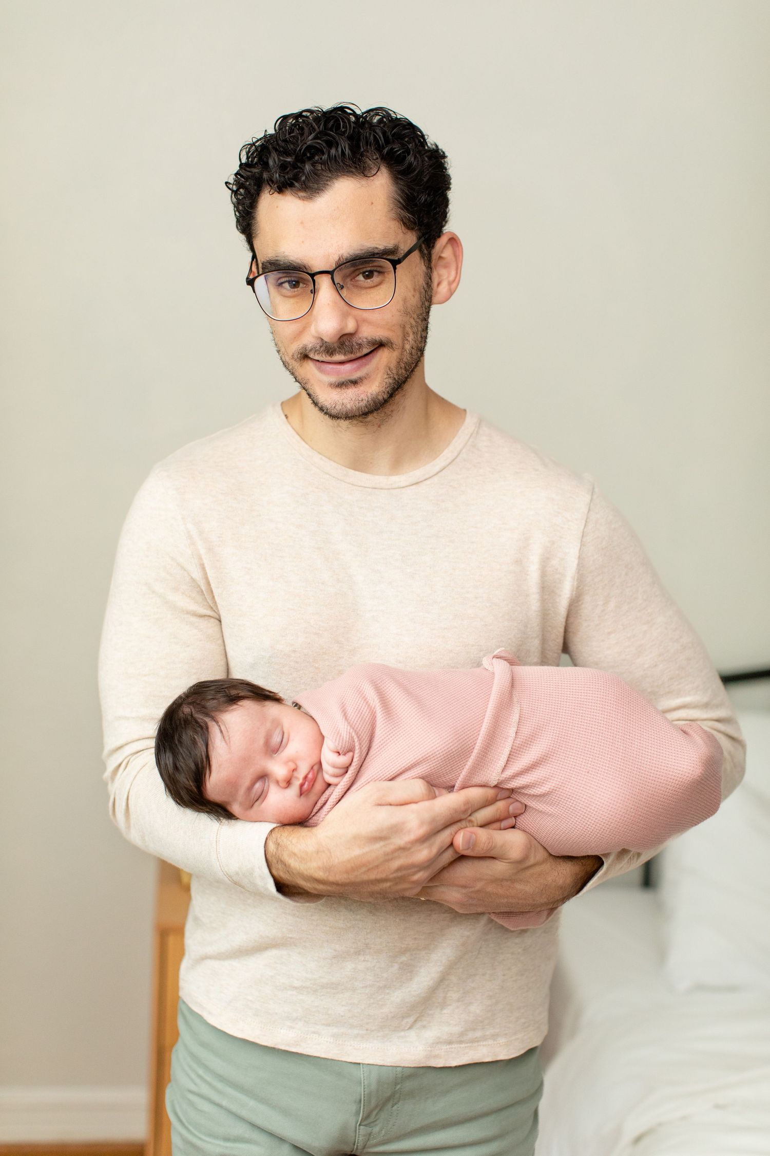 Parent wearing glasses smiles while holding sleeping newborn in pink swaddle.