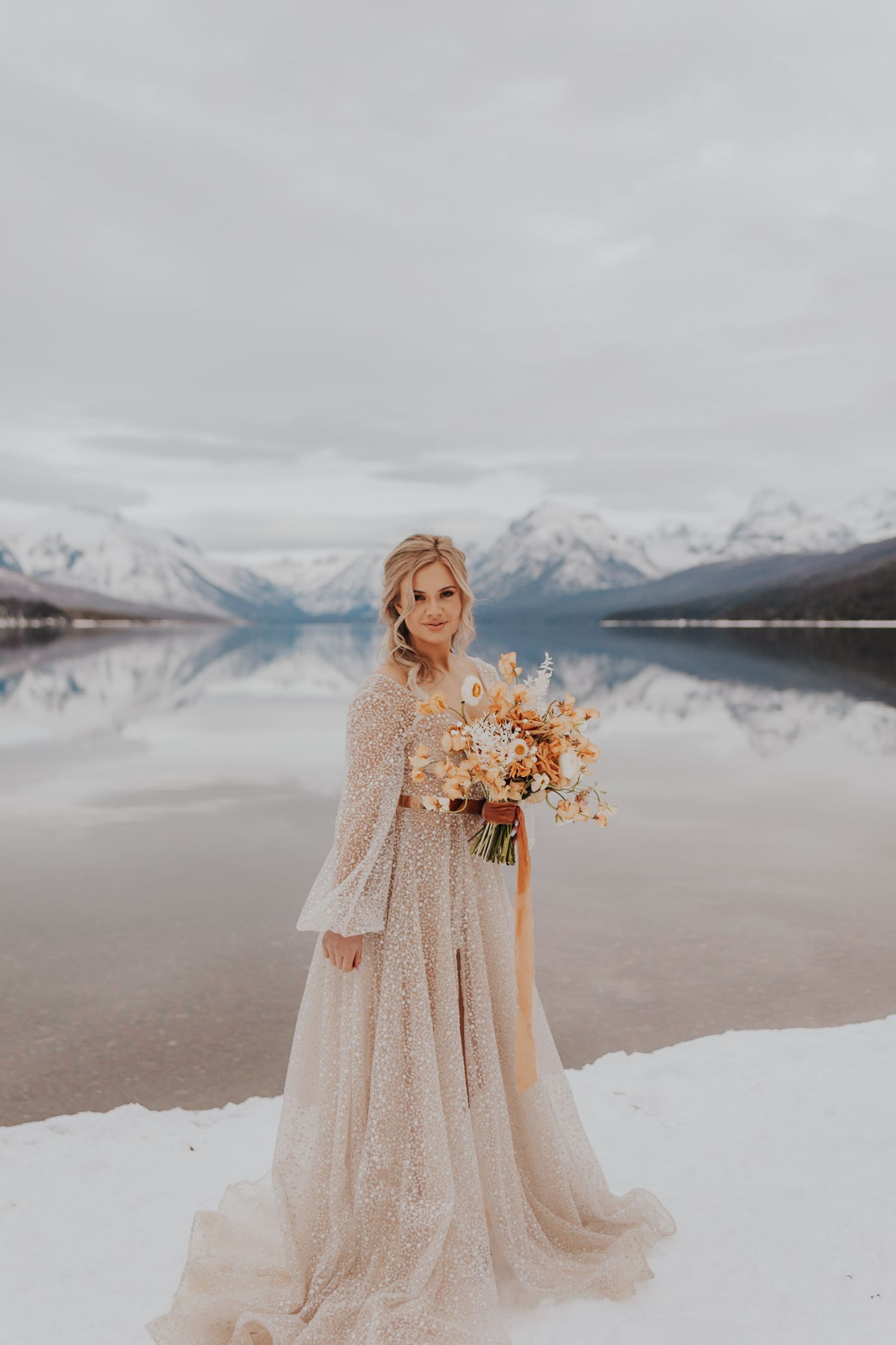 Bride stands on the edge of a lake in Glacier National Park with a sparkly wedding dress and orange accent bouquet