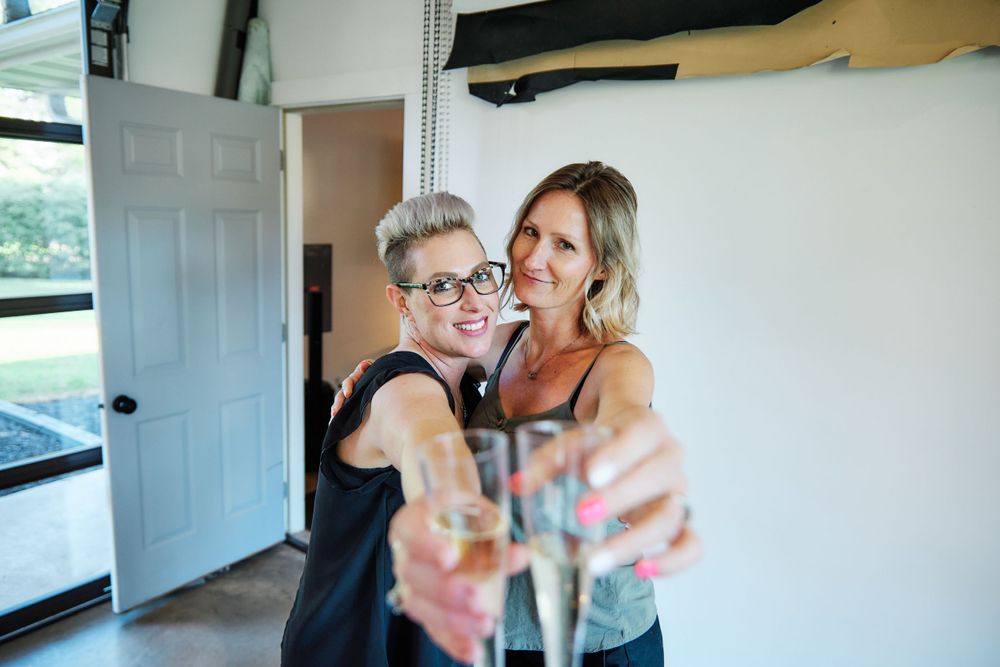 Two people toasting colorful drinks together while smiling in a casual indoor setting.