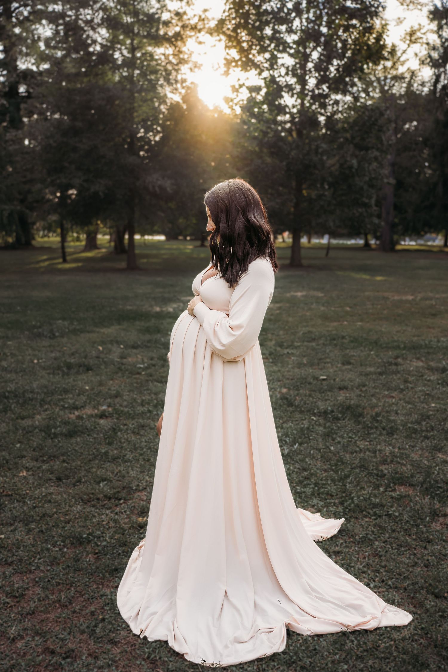 Photograph of a pregnant Delaware Mom dressed in a cream colored maternity gown holding her belly, while looking down.