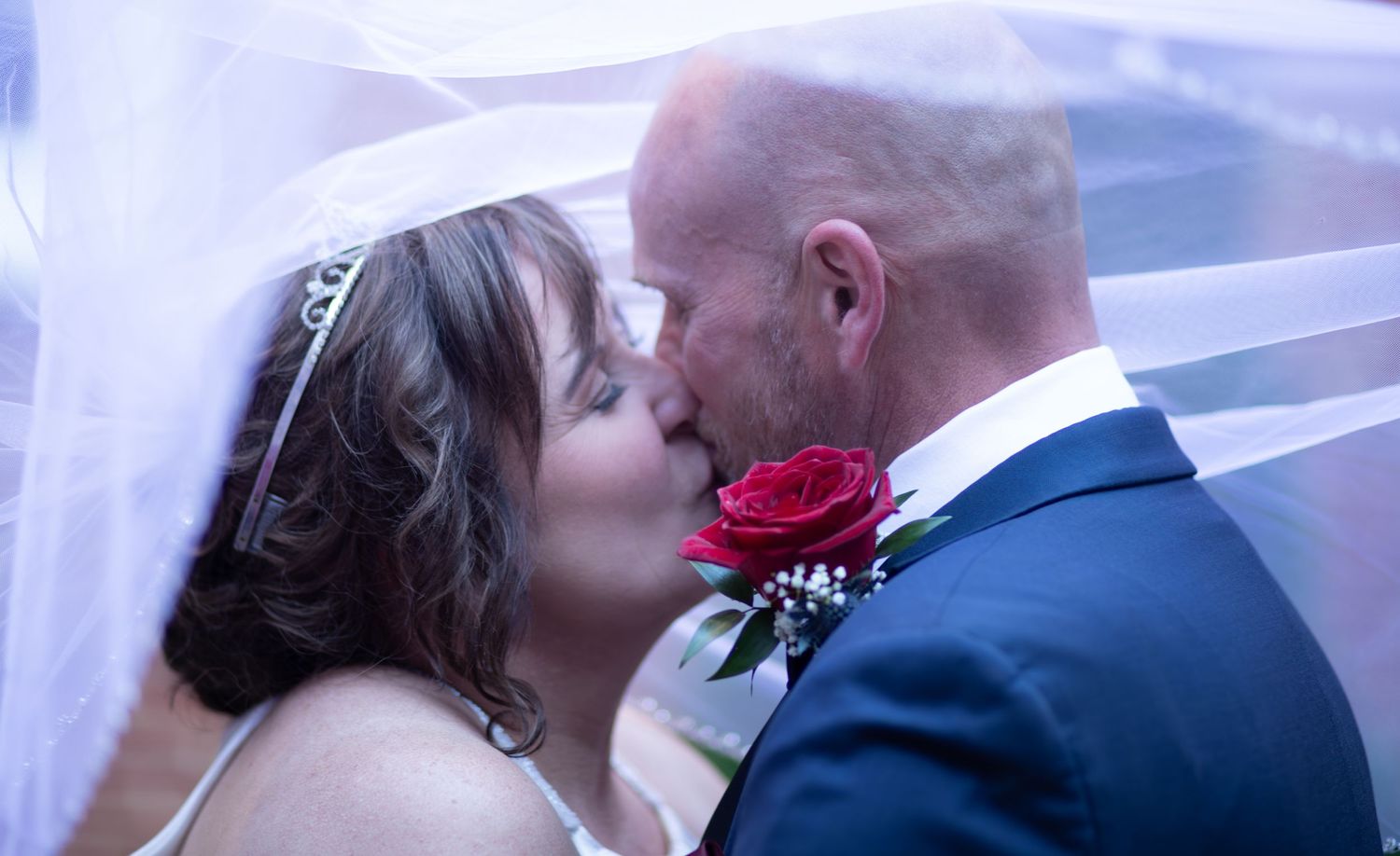 A romantic wedding moment with a bride and groom embracing, featuring a red rose boutonniere.