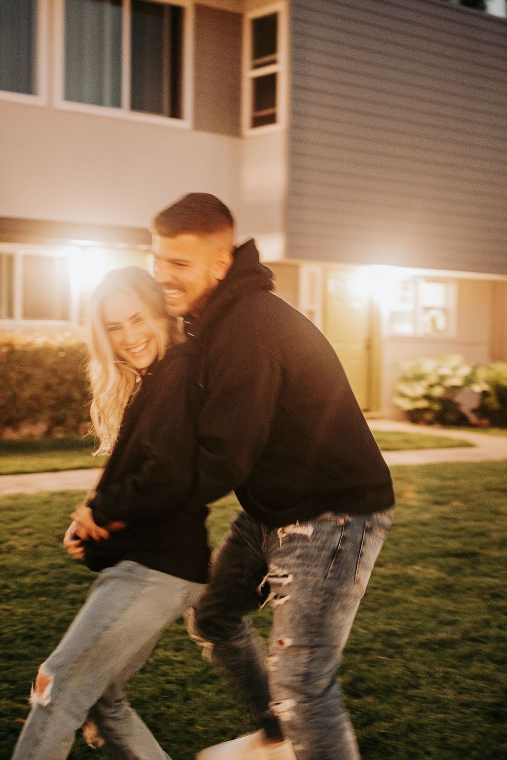 Two people playfully chase each other on a lawn at sunset with warm lighting.