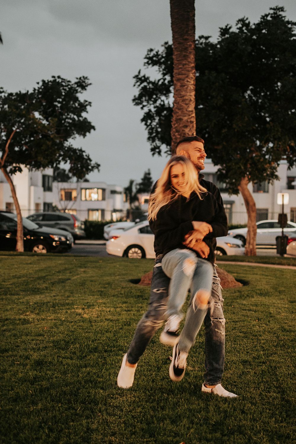 A couple shares a fun moment dancing and playing on grass near palm trees.