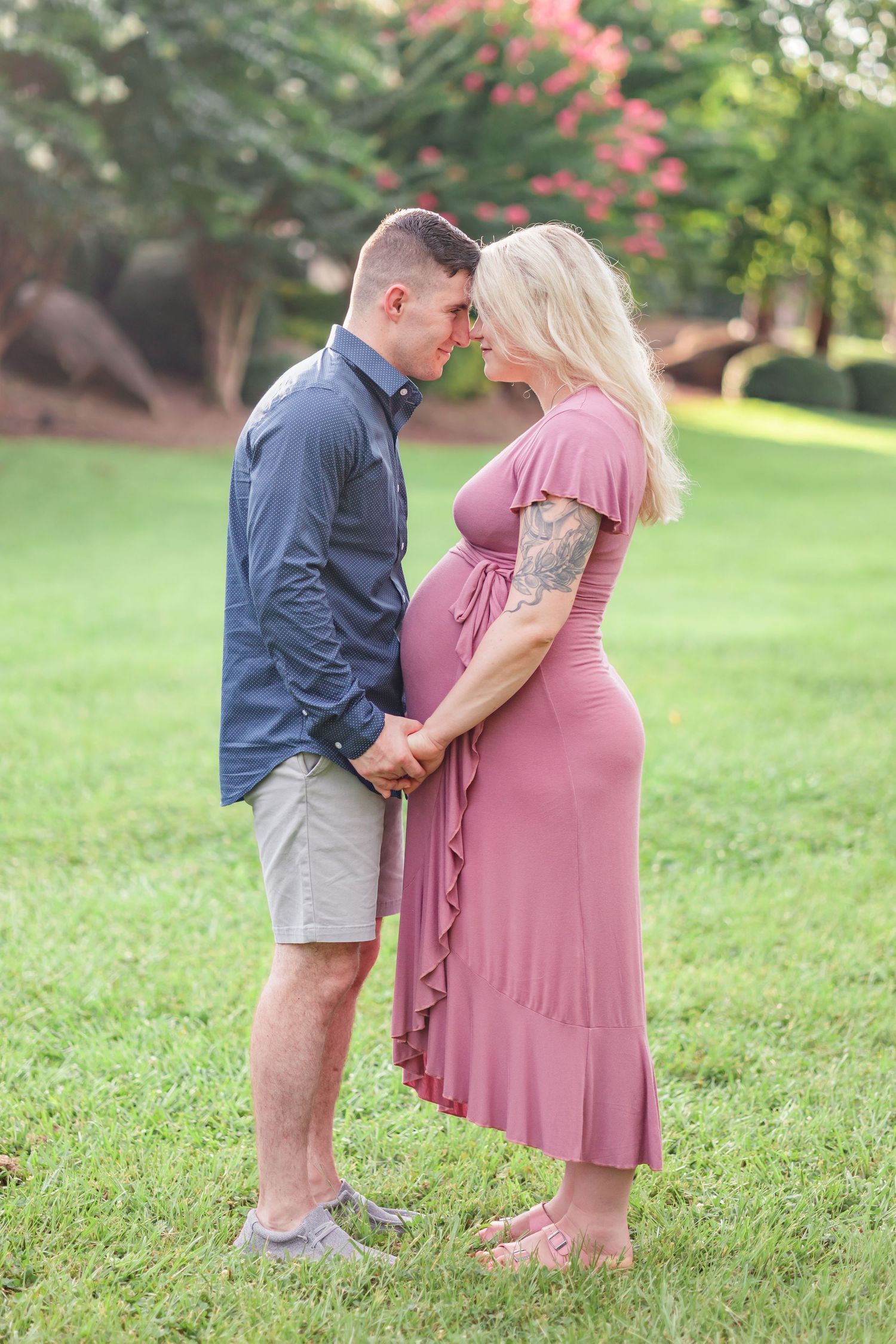 Expectant couple shares an intimate moment outdoors in a garden setting, wearing blue and pink outfits.