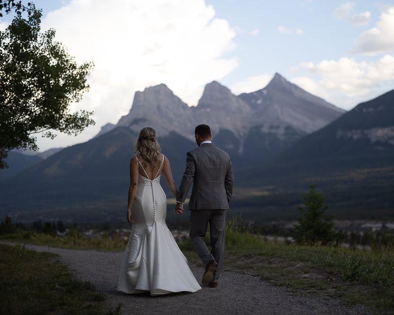 Wedding couple walks mountain path with Three Sisters peaks in background.