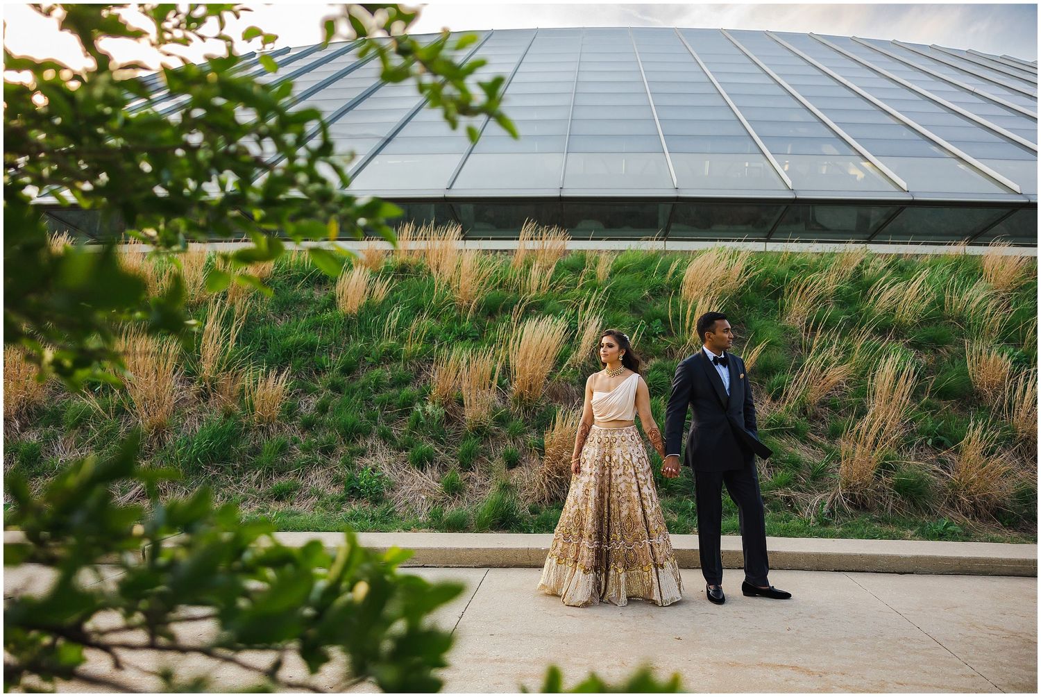 Artistic shot through foliage of couple standing by glass building and natural landscaping.
